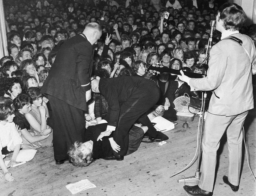 Сопровождающие выносят со сцены девушку, которая упала в обморок, наблюдая за пением Джона Леннона