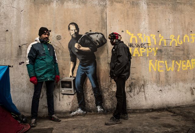 Британский уличный художник Бэнкси изобразил бывшего главу корпорации Apple Стива Джобса в образе мигранта из Сирии. Граффити появилось на стене лагеря беженцев во французском Кале. В одной руке Джобс несет компьютер, в другой – наплечный мешок с вещами. Рядом с граффити на стене находится надпись: "Никто не заслуживает жить такой жизнью". Фото: AFP